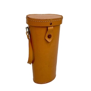 May include: A cylindrical, tan leather case with a matching lid. The case has a stitched seam around the top and bottom, with a small strap and button closure. A small handle is attached to the side with a metal clasp.