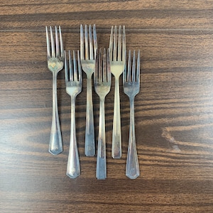 May include: Six silver forks with ornate handles. The forks are arranged in a row on a wooden surface.