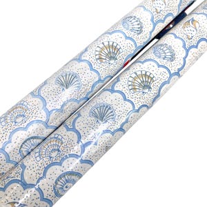 May include: Two rolls of wrapping paper featuring a repeating pattern of seashells in shades of blue and gold. The design includes a scalloped blue border against a white background with small gold dots, creating a nautical aesthetic on the paper.