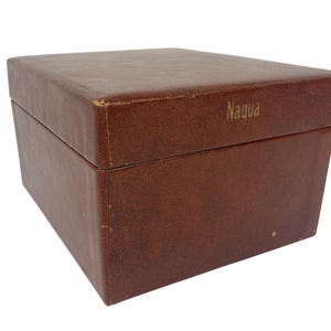 Puede incluir: Una caja rectangular marrón con una superficie texturizada. La palabra "Naqua" está impresa en dorado en la parte superior de la caja. La caja está cerrada y parece estar hecha de cuero o un material similar.