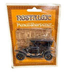 Puede incluir: Un sacapuntas de estilo vintage en un paquete de plástico transparente. El sacapuntas es una réplica en miniatura de un coche negro clásico. El embalaje es naranja con las palabras "NOSTALGIC Pencil Sharpener".