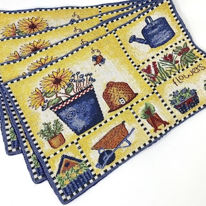 May include: A set of four decorative placemats with a yellow background and a blue border. Each placemat features a repeating pattern of garden-themed squares with sunflowers, watering cans, and the word "flowers".