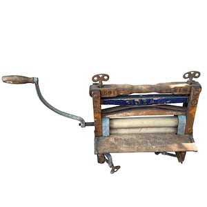 Puede incluir: Antiguo escurridor de ropa de madera con manivela. El escurridor tiene un marco de madera con rodillos y herrajes metálicos. La parte superior tiene detalles pintados en azul y blanco. El texto "HORSE SHOE BRAND CLOTHES WRINGER" es visible.