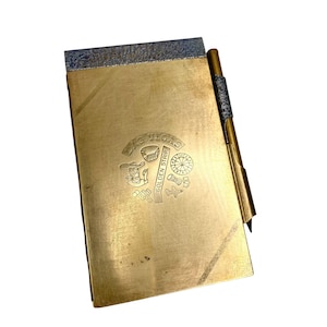 May include: A rectangular brass notepad holder with a matching pen attached to the side. The front surface features an engraved design with the text LAS VEGAS GOLDEN STRIP, a cowboy figure, a roulette wheel, dice, and an airplane icon.