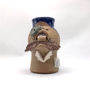 Puede incluir: Un jarrón de cerámica con forma de persona con un sombrero azul, barba marrón y un solo ojo. El jarrón tiene un esmalte marrón y azul y una etiqueta blanca con la letra "M" en ella.