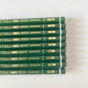 May include: A close-up of a stack of green pencils with gold lettering. The pencils are labeled "STELL 9000 GERMANY" and marked with "HB" or "H". The pencils are arranged in a neat, parallel row.