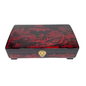 May include: Black rectangular box with a red Asian landscape design on the lid and sides. The box features a gold-colored clasp and is elevated on small feet. The design includes mountains, buildings, and bridges.
