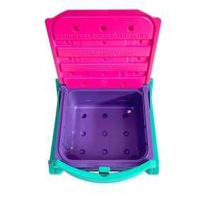 Puede incluir: Una nevera de pl&aacute;stico rosa y morada con un asa turquesa. La tapa est&aacute; abierta y el interior es morado. La tapa tiene el texto "DRAIN WATER BEFORE TRANSPORTING FREEZE WATER BOTTLE PROVIDED (OPTIONAL)"