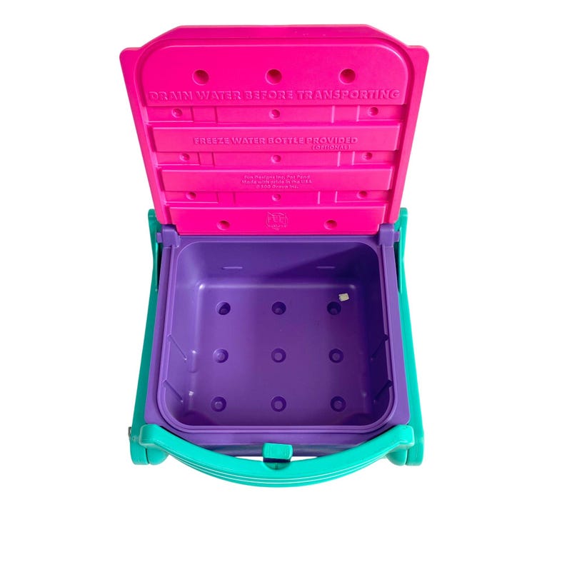 Puede incluir: Una nevera de pl&aacute;stico rosa y morada con un asa turquesa. La tapa est&aacute; abierta y el interior es morado. La tapa tiene el texto "DRAIN WATER BEFORE TRANSPORTING FREEZE WATER BOTTLE PROVIDED (OPTIONAL)"