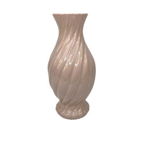 Könnte beinhalten: Eine hellrosa Keramikvase mit einem gedrehten, gerippten Design. Die Vase hat eine ausgestellte Oberseite und einen abgerundeten Boden. Die glänzende Oberfläche reflektiert das Licht und hebt das Spiralmuster hervor. Diese dekorative Vase eignet sich für Blumen oder als eigenständiges Akzentstück.