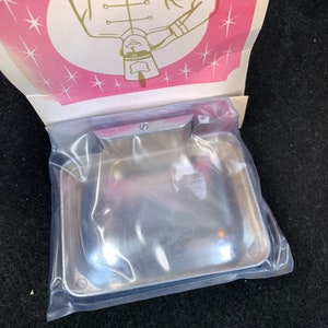 May include: A new, shiny, rectangular metal ashtray is sealed in a clear plastic bag. The ashtray has rounded corners and a small, curved handle. The packaging includes a pink and white decorative top with a cartoon illustration.