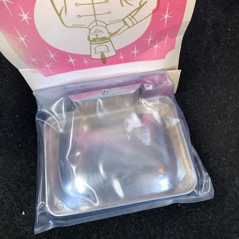 May include: A new, shiny, rectangular metal ashtray is sealed in a clear plastic bag. The ashtray has rounded corners and a small, curved handle. The packaging includes a pink and white decorative top with a cartoon illustration.