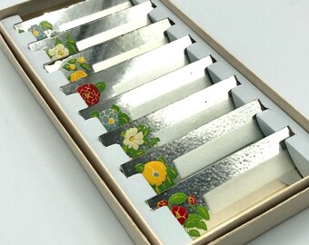 May include: A set of eight silver place card holders with floral designs. Each holder features a different floral pattern with yellow, red, blue, and green accents.