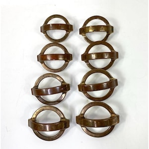 Puede incluir: Ocho anillos metálicos antiguos de color bronce con un diseño circular y una barra plana y rectangular en el centro. Los anillos están dispuestos en dos columnas de cuatro, mostrando su pátina envejecida y estética vintage.