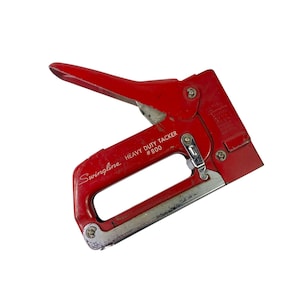 Puede incluir: Una grapadora manual roja Swingline Heavy Duty Tacker #800. La grapadora es roja con detalles plateados y presenta el logotipo de Swingline y el número de modelo. El mango está abierto, listo para usar.