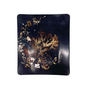 Könnte beinhalten: Ein rechteckiges, dunkelblaues Dekotablett mit abgerundeten Ecken. Es zeigt ein detailliertes, goldbraunes botanisches Design in der Mitte, mit verstreuten Gold- und Weißakzenten. Das Design erinnert an eine stilisierte Pflanze oder Koralle.