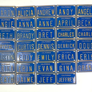Puede incluir: Un conjunto de 25 placas de matrícula azules y doradas con diferentes nombres impresos en ellas. Cada placa tiene la palabra "California" impresa en la parte superior.