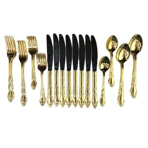 May include: A collection of gold-colored flatware, including forks, knives, and spoons, arranged on a white surface. The knives have black blades. The handles of the forks, knives, and spoons have a decorative, ornate design.
