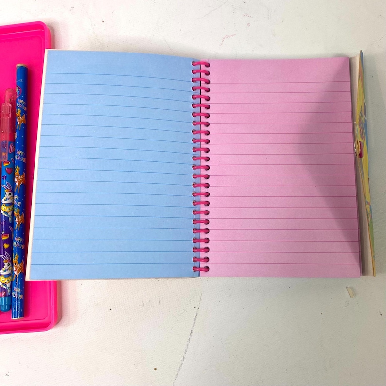 May include: Open notebook with blue and pink lined pages, bound by a pink spiral. A pink plastic tray with blue and purple pencils is visible on the left. The notebook is open on a white surface.