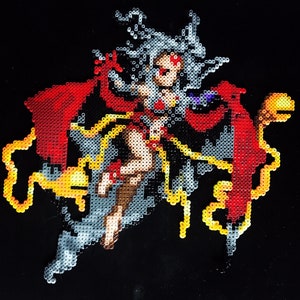 May include: A beaded figure of a woman with long white hair, wearing a red and black cape, and holding a golden staff. The figure is surrounded by yellow beads.