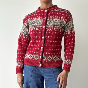 May include: A red cardigan sweater with a geometric pattern in white, gray, and black. The sweater has a button-up front and is paired with blue denim jeans. The sweater's design features diamond and dot patterns.
