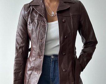 Può includere: Giacca in pelle marrone scuro con colletto e bottoni. La giacca presenta una vestibilità sartoriale con dettagli di cucitura verticali e una cintura. Le maniche sono lunghe e la giacca è indossata sopra un top bianco e jeans blu.