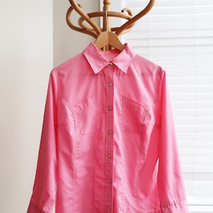 Puede incluir: Una camisa rosa de manga larga con botones, cuello y dos bolsillos. La camisa está colgada en un perchero de madera.