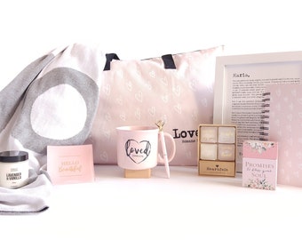 May include: A pink and white gift basket with a blanket, mug, candle, card, and a book. The mug says "Loved" and the book says "Promises for Your Soul".