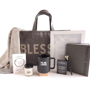 May include: A gift basket with a brown tote bag that says "Bless" on it. The basket also includes a blanket, a candle, a mug that says "You Are Loved", a prayer journal, a book of bible promises, a pen, and a bracelet.