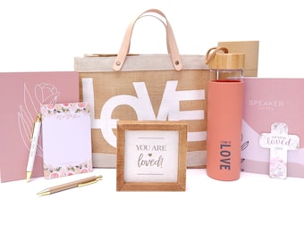 May include: A gift basket with a pink and white floral theme. It includes a tote bag with the word "LOVE" printed on it, a pink water bottle with the word "LOVE" printed on it, a pink notebook, a notepad with floral designs, two pens, a wooden sign with the words "You are loved!" and a bookmark with a cross and the words "You are loved" printed on it.