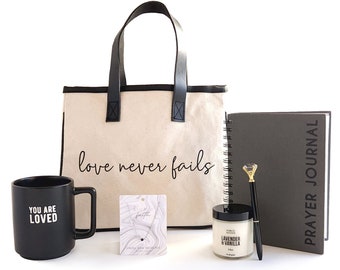 May include: A gift set with a black tote bag that says "love never fails", a black mug that says "you are loved", a black and gray prayer journal, a small candle, and a gold pen.