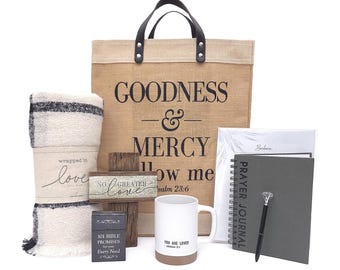 May include: A collection of inspirational gifts, including a burlap tote bag with "GOODNESS & MERCY" text, a blanket, a wooden cross, a mug, a journal, and a pen. The mug reads "YOU ARE LOVED". The blanket is cream and black.