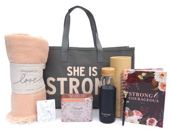 May include: A gift set featuring a gray tote bag with "SHE IS STRONG" text, a peach-colored blanket, a water bottle, a journal, a pen, and a decorative tin. The blanket is wrapped with a label that says "wrapped in love".