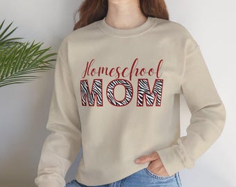 Puede incluir: Una sudadera beige de cuello redondo con el texto Homeschool en letra cursiva roja sobre la palabra MOM en letras de bloque, rellena con un patrón de rayas de cebra en blanco y negro y un borde rojo.