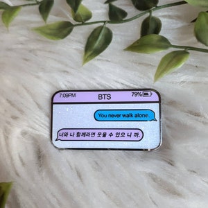 May include: A glittery enamel pin featuring a text message bubble with the text "You never walk alone." and the Korean text "너와 나 함께라면 웃을 수 있으니까."  The pin also includes a text message bar with the text "BTS" and a battery icon with 79% battery life.
