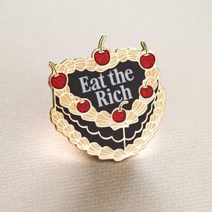 May include: A gold-coloured pin badge shaped like a layered cake, adorned with red cherries. The cake features a black centre with the white text "Eat the Rich". Cream-coloured frosting decorates the cake.