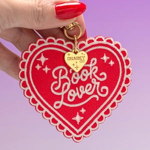 Puede incluir: Un parche bordado rojo en forma de corazón con un borde festoneado blanco. El texto "Book lover" está bordado en cursiva blanca, acentuado con pequeños iconos de estrellas blancas. Un dije de metal dorado grabado con "SNARKY CO." cuelga de un broche dorado.