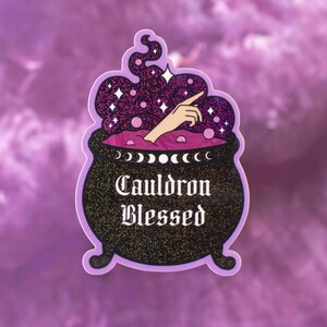 Puede incluir: Una pegatina con un caldero negro con el texto "Cauldron Blessed". El caldero tiene un borde morado y está lleno de una poción morada brillante. Una mano emerge de la poción, apuntando hacia arriba. El fondo es un degradado de morado.