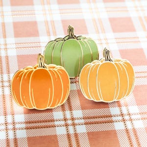 May include: Three pumpkin-shaped enamel pins in shades of orange and green with gold-colored outlines and stems. The pins are arranged on a plaid patterned surface, evoking a fall or harvest theme.