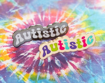 May include: Two enamel pins with the word "Autistic" in colorful, bubble-like letters. The top pin is black with white letters, and the bottom pin is gold with purple, green, pink, and blue letters.
