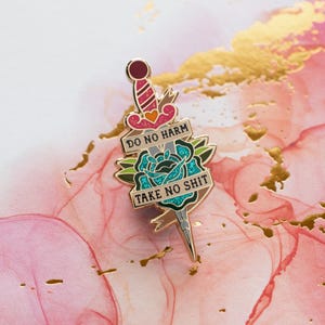 May include: A gold-colored enamel pin featuring a dagger piercing a teal rose. The pin includes the phrases "DO NO HARM" and "TAKE NO SHIT" on banners. The pin has red, pink, and green accents, set against a pink and gold background.