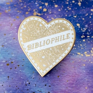 Puede incluir: Pin de esmalte en forma de corazón dorado con detalles en esmalte blanco. El pin presenta un diseño decorativo de libros, flores y estrellas. La palabra "Bibliophile" está escrita en blanco en el centro del pin.