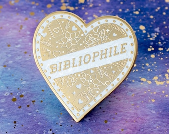 May include: Gold heart-shaped enamel pin with white enamel detailing. The pin features a decorative design of books, flowers, and stars. The word "Bibliophile" is written in white across the center of the pin.