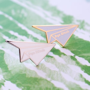 May include: Two paper airplane pins with the text "Bye Bitch" in gold lettering. The pins are silver and one is white and the other is light grey.