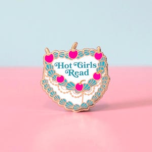 May include: A gold-toned metal enamel pin shaped like a cake with white frosting, teal decorative piping, and bright pink cherries. The center of the cake features the text Hot Girls Read in teal lettering. The pin has a scalloped edge design.