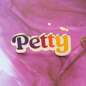 May include: A gold-colored enamel pin with the word "Petty" in a retro font. The letters are in shades of purple, red, orange, and yellow. The pin is set against a pink and purple background.