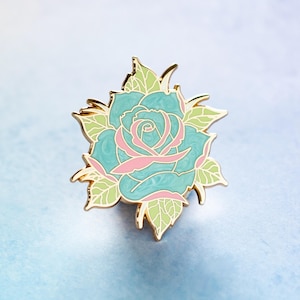 May include: A teal and pink rose enamel pin with gold-colored trim. The pin features a detailed design of a rose with green leaves. The rose is a vibrant teal color with pink accents. The leaves are light green.