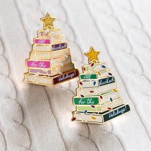 May include: Two gold-coloured enamel pins shaped like Christmas trees made of books. Each tree has a gold star on top and is decorated with a string of lights. The books have the phrases "All booked for the Holidays!" on them.