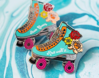 Könnte beinhalten: Zwei türkisfarbene Rollschuh-Anstecknadeln mit orangefarbenen Akzenten, rosa Rädern und floralen Verzierungen. Auf einer Nadel steht "Skate Queen", auf der anderen "Roll with it...". Beide Nadeln haben Glitzerakzente.