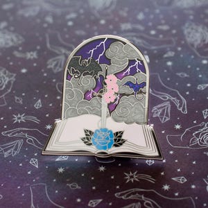 Peut inclure: Une épingle en émail argentée représentant un livre avec une épée qui le traverse. Le livre est ouvert et une rose bleue est sur la page. L'arrière-plan est un ciel orageux avec des éclairs et deux chauves-souris.